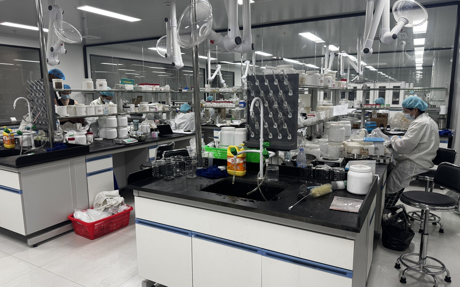 Close lab bench scene with several protected staff members, raw materials and glassware in use