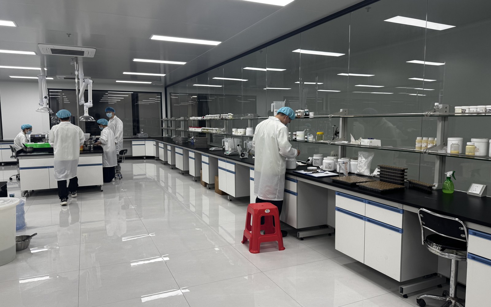 Wide R&D lab view with multiple protected staff members working at different formulation benches